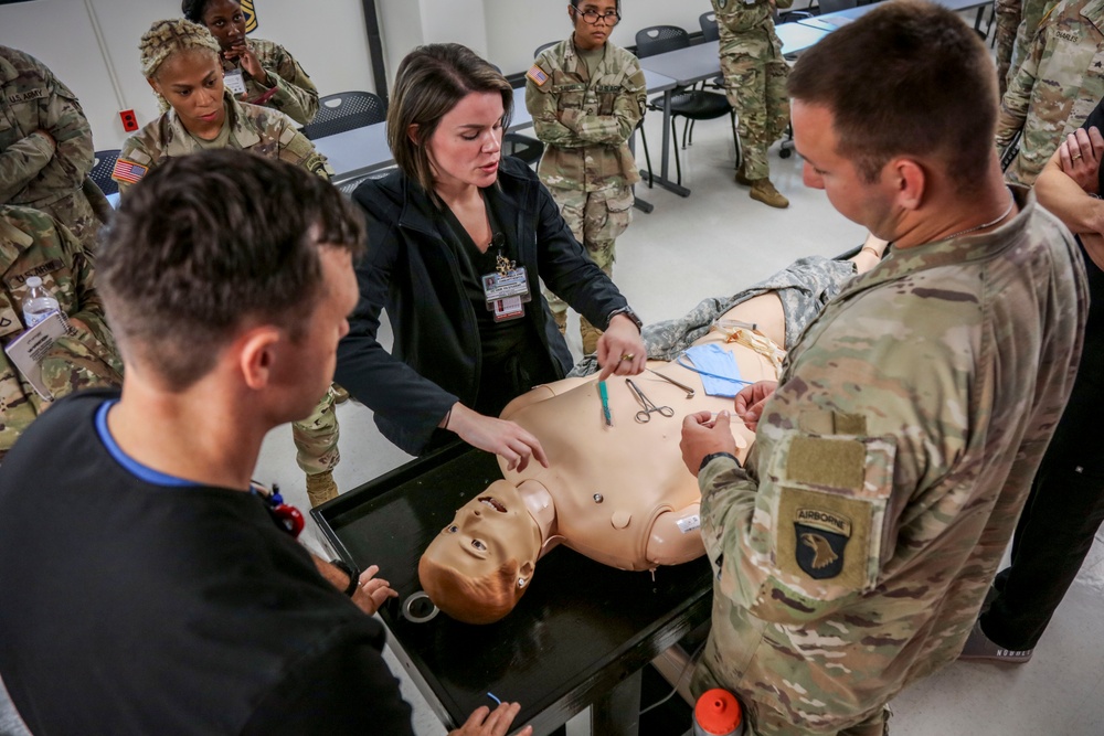 A civilian medical instructor is conducting a hands-on medical training session for U.S. Army soldiers in a classroom setting. The instructor and a soldier wearing a 101st Airborne Division patch are demonstrating a medical procedure on a anatomical training mannequin. Multiple soldiers in Army Combat Uniforms observe the demonstration from around the table.