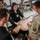 A civilian medical instructor is conducting a hands-on medical training session for U.S. Army soldiers in a classroom setting. The instructor and a soldier wearing a 101st Airborne Division patch are demonstrating a medical procedure on a anatomical training mannequin. Multiple soldiers in Army Combat Uniforms observe the demonstration from around the table.