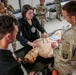 A civilian medical instructor is conducting a hands-on medical training session for U.S. Army soldiers in a classroom setting. The instructor and a soldier wearing a 101st Airborne Division patch are demonstrating a medical procedure on a anatomical training mannequin. Multiple soldiers in Army Combat Uniforms observe the demonstration from around the table.