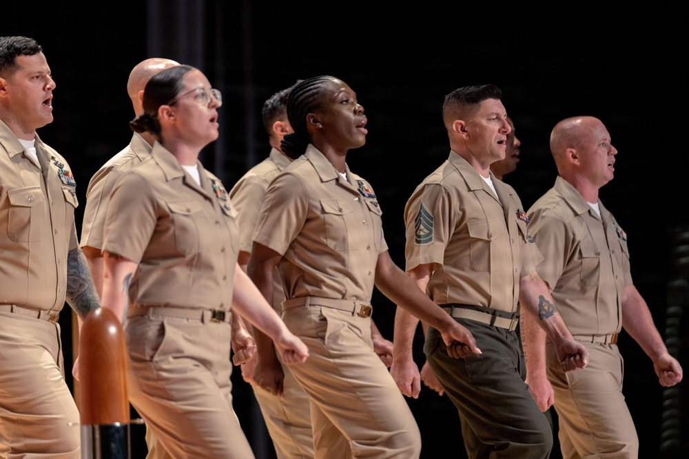 A group of six military personnel in khaki/tan service uniforms are standing in a line on a stage, appearing to sing or recite something in unison. The group includes members from multiple branches of the U.S. military, identifiable by differing uniform details and insignia. They appear to be participating in a formal ceremony or performance.