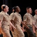 A group of six military personnel in khaki/tan service uniforms are standing in a line on a stage, appearing to sing or recite something in unison. The group includes members from multiple branches of the U.S. military, identifiable by differing uniform details and insignia. They appear to be participating in a formal ceremony or performance.