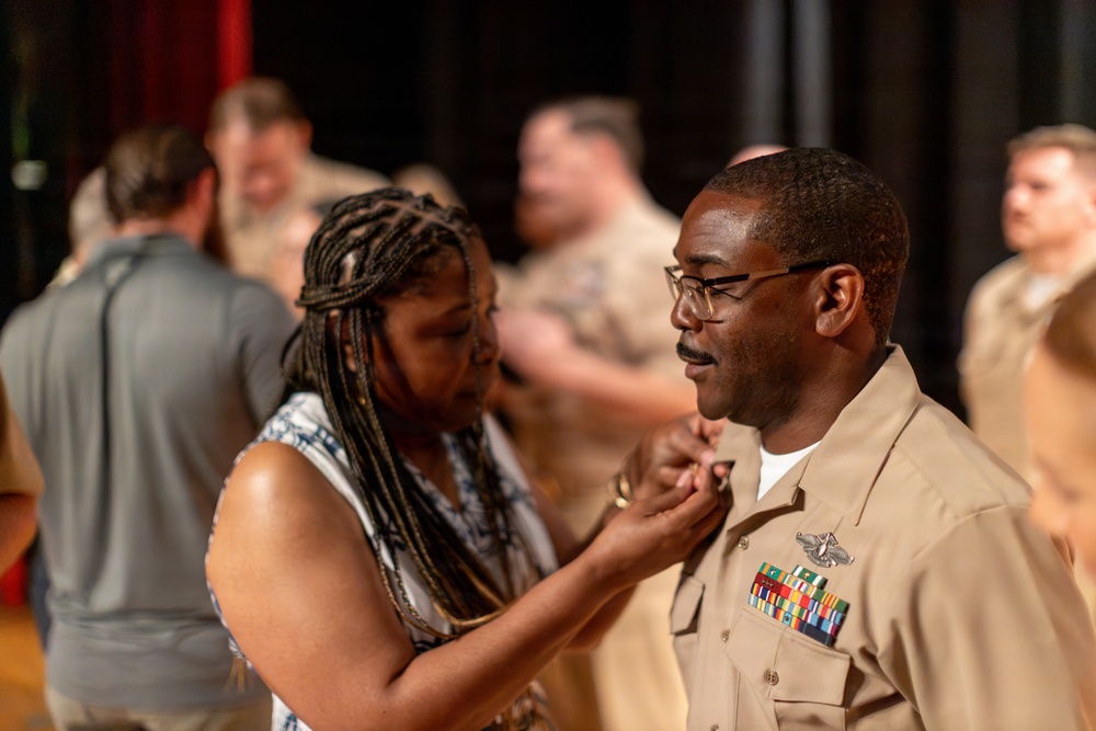 A woman is pinning something onto the uniform of a male U.S. Navy service member during what appears to be a military promotion or award ceremony. The sailor is wearing a tan/khaki service uniform adorned with multiple ribbons, a warfare device, and other insignia. Several other uniformed personnel and civilians are visible in the blurred background, suggesting a group ceremony event.