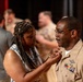 A woman is pinning something onto the uniform of a male U.S. Navy service member during what appears to be a military promotion or award ceremony. The sailor is wearing a tan/khaki service uniform adorned with multiple ribbons, a warfare device, and other insignia. Several other uniformed personnel and civilians are visible in the blurred background, suggesting a group ceremony event.