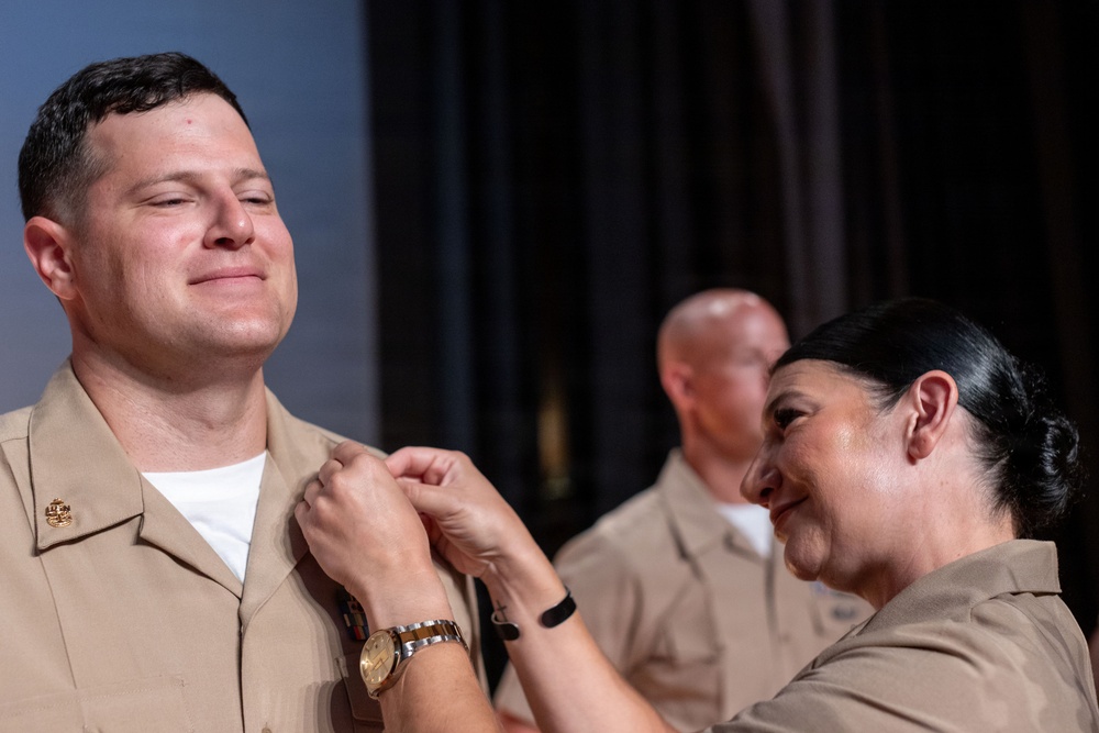 A military promotion or pinning ceremony is taking place, with a woman in U.S. Navy Service Uniform (NWU Type III/khaki) pinning a device or insignia onto the collar of a male sailor who is smiling with eyes closed. The emotional moment suggests a significant milestone, likely a promotion to Chief Petty Officer or similar rank. A third uniformed individual is visible in the blurred background.