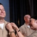 A military promotion or pinning ceremony is taking place, with a woman in U.S. Navy Service Uniform (NWU Type III/khaki) pinning a device or insignia onto the collar of a male sailor who is smiling with eyes closed. The emotional moment suggests a significant milestone, likely a promotion to Chief Petty Officer or similar rank. A third uniformed individual is visible in the blurred background.