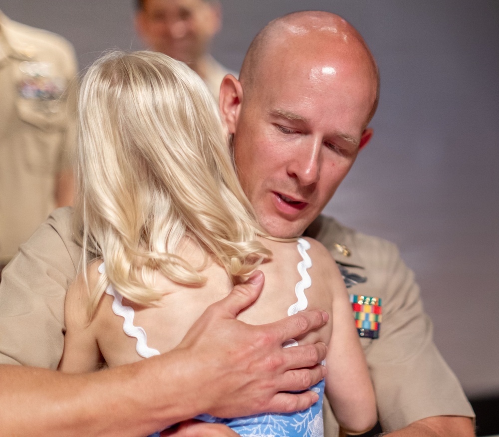 A bald military serviceman in a tan/khaki uniform adorned with service ribbons is embracing a young blonde child. The moment appears emotionally significant, possibly a reunion or ceremony. A second uniformed individual is visible but out of focus in the background.
