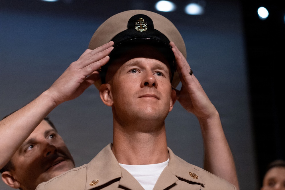 A military ceremony appears to be taking place in which a chief petty officer's cover (hat) is being placed on a service member's head by others standing behind him. The central figure is wearing a tan/khaki U.S. Navy uniform with gold insignia, and the black-brimmed cover features a gold chief petty officer anchor device. This is consistent with a U.S. Navy Chief Petty Officer pinning or frocking ceremony.