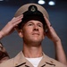 A military ceremony appears to be taking place in which a chief petty officer's cover (hat) is being placed on a service member's head by others standing behind him. The central figure is wearing a tan/khaki U.S. Navy uniform with gold insignia, and the black-brimmed cover features a gold chief petty officer anchor device. This is consistent with a U.S. Navy Chief Petty Officer pinning or frocking ceremony.