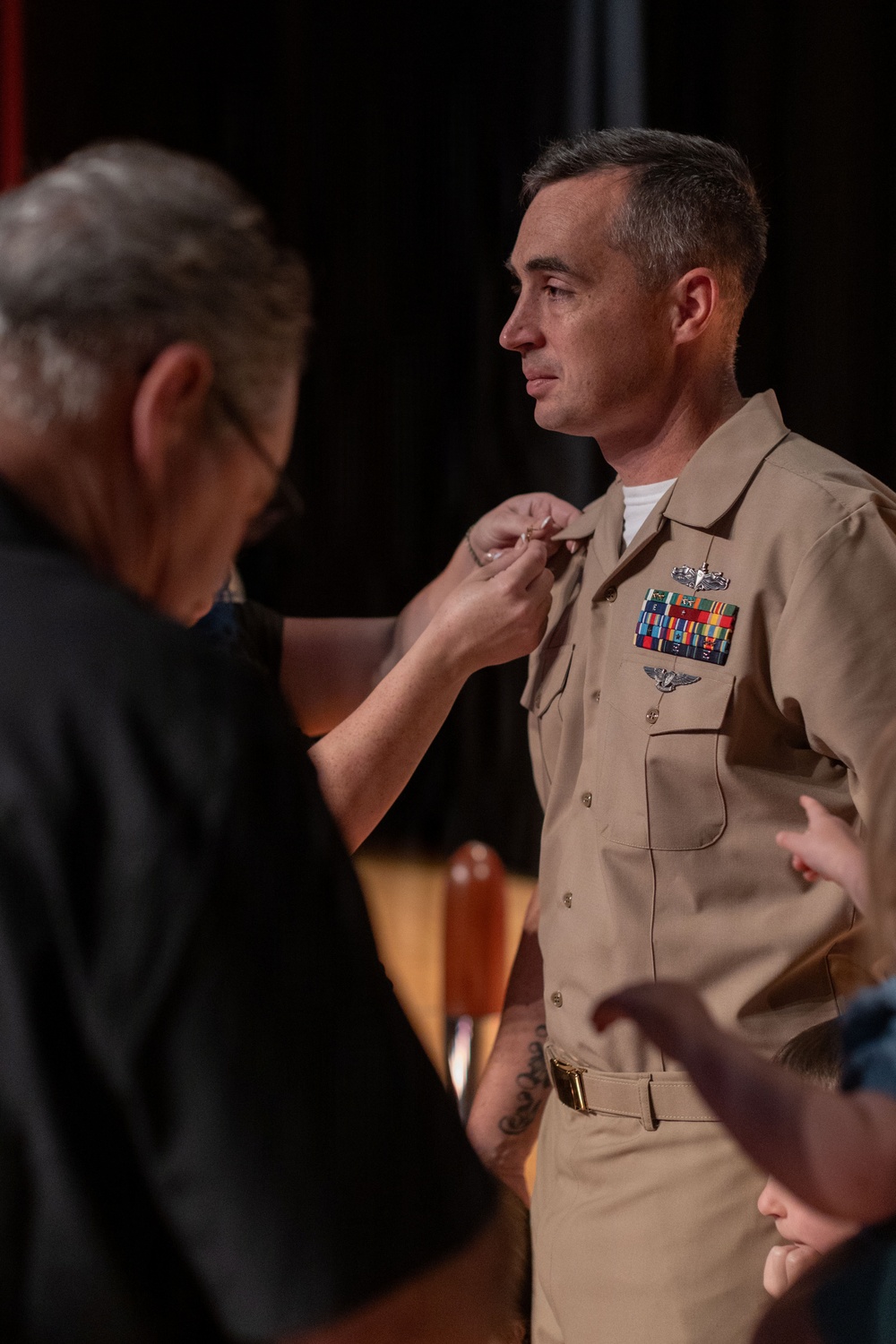 A U.S. Navy officer in khaki service uniform is having an insignia or device pinned onto his collar during what appears to be a formal military ceremony. A civilian in a black shirt is performing the pinning, suggesting this may be a promotion or qualification ceremony. The officer displays multiple service ribbons, aviation wings, and other military badges on his uniform.