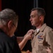 A U.S. Navy officer in khaki service uniform is having an insignia or device pinned onto his collar during what appears to be a formal military ceremony. A civilian in a black shirt is performing the pinning, suggesting this may be a promotion or qualification ceremony. The officer displays multiple service ribbons, aviation wings, and other military badges on his uniform.