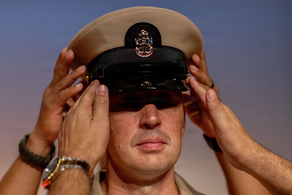 A U.S. Navy Chief Petty Officer is having his combination cover (cap) placed on his head, likely during a promotion or pinning ceremony. Two sets of hands are visible adjusting the khaki cover, which bears the USN anchor insignia. This appears to be a formal military advancement ritual.