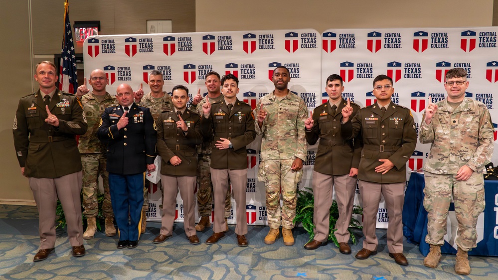 A group of approximately 12 military personnel pose for a formal group photo in front of a Central Texas College branded backdrop. Most individuals are holding up one finger, suggesting a '#1' or celebratory gesture. The group includes personnel in various U.S. Army uniforms including dress greens, Army Combat Uniforms (ACU/OCP), and Class A uniforms.