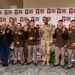 A group of approximately 12 military personnel pose for a formal group photo in front of a Central Texas College branded backdrop. Most individuals are holding up one finger, suggesting a '#1' or celebratory gesture. The group includes personnel in various U.S. Army uniforms including dress greens, Army Combat Uniforms (ACU/OCP), and Class A uniforms.