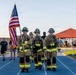 Three firefighters in full turnout gear and helmets march in formation on a blue running track while carrying an American flag. The event appears to be a memorial or tribute run, likely a 9/11 remembrance ceremony, with other participants walking and running in the background. The setting includes an outdoor athletic facility with a red-roofed structure visible in the background.