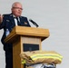 An older man in a dark navy dress uniform with rank insignia and medals is speaking at a wooden podium with a microphone. Firefighter turnout gear with yellow reflective stripes is displayed in the foreground, suggesting a fire service ceremony or memorial event. A projection screen is visible in the background.