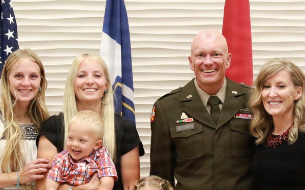A group of eight people, including a U.S. Army officer in dress uniform, pose together in what appears to be a military ceremony or promotion event. The group includes adults, a young boy being held, and a young girl in front, suggesting a family gathering. Three flags are visible in the background, including the U.S. flag and what appears to be Army-related flags.