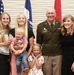 A group of eight people, including a U.S. Army officer in dress uniform, pose together in what appears to be a military ceremony or promotion event. The group includes adults, a young boy being held, and a young girl in front, suggesting a family gathering. Three flags are visible in the background, including the U.S. flag and what appears to be Army-related flags.
