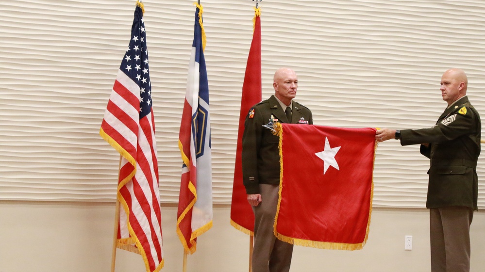 Two U.S. Army soldiers are participating in a formal military ceremony, likely a promotion or change of command. One soldier holds a red brigadier general flag (one white star), while a senior NCO assists in presenting or unfurling it. Three flags are visible in the background: the U.S. flag, what appears to be a unit or state flag, and a red general officer flag.