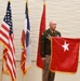 Two U.S. Army soldiers are participating in a formal military ceremony, likely a promotion or change of command. One soldier holds a red brigadier general flag (one white star), while a senior NCO assists in presenting or unfurling it. Three flags are visible in the background: the U.S. flag, what appears to be a unit or state flag, and a red general officer flag.