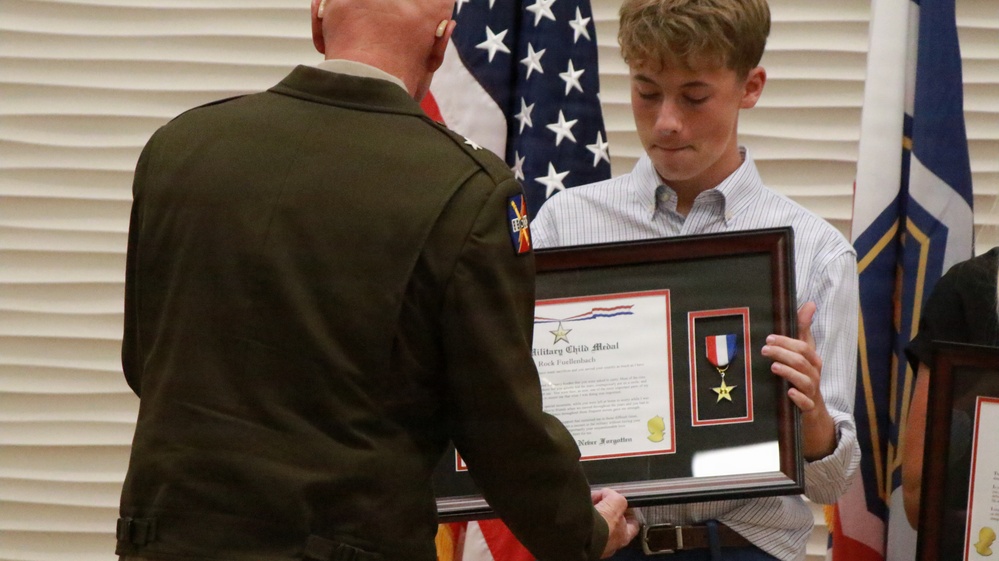 A military officer in dress uniform is presenting a framed 'Military Child Medal' award to a young civilian male during a formal ceremony. The framed display contains a certificate and a medal with red, white, and blue ribbon. American flags and other military flags are visible in the background.