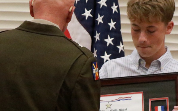 A military officer in dress uniform is presenting a framed 'Military Child Medal' award to a young civilian male during a formal ceremony. The framed display contains a certificate and a medal with red, white, and blue ribbon. American flags and other military flags are visible in the background.