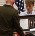 A military officer in dress uniform is presenting a framed 'Military Child Medal' award to a young civilian male during a formal ceremony. The framed display contains a certificate and a medal with red, white, and blue ribbon. American flags and other military flags are visible in the background.
