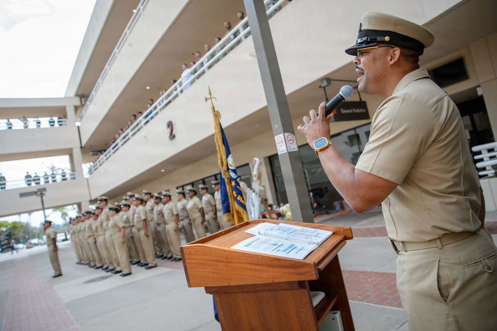 A senior U.S. Navy enlisted officer in khaki service uniform speaks into a microphone at a wooden podium during an outdoor military ceremony. A large formation of sailors in matching khaki uniforms stands at attention in the background. The event appears to take place at a military installation courtyard with observers watching from an upper-level balcony.