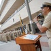 A senior U.S. Navy enlisted officer in khaki service uniform speaks into a microphone at a wooden podium during an outdoor military ceremony. A large formation of sailors in matching khaki uniforms stands at attention in the background. The event appears to take place at a military installation courtyard with observers watching from an upper-level balcony.