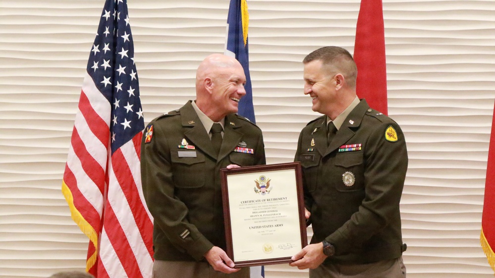 Two U.S. Army officers in green service uniforms are smiling and jointly holding a framed Certificate of Retirement during what appears to be a military retirement ceremony. The certificate identifies the retiree as a Brigadier General in the United States Army. The ceremony is taking place in front of multiple flags including the American flag and what appear to be unit or state flags.