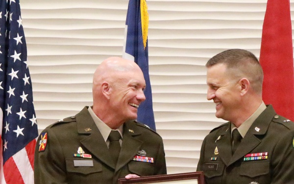 Two U.S. Army officers in green service uniforms are smiling and jointly holding a framed Certificate of Retirement during what appears to be a military retirement ceremony. The certificate identifies the retiree as a Brigadier General in the United States Army. The ceremony is taking place in front of multiple flags including the American flag and what appear to be unit or state flags.
