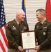 Two U.S. Army officers in green service uniforms are smiling and jointly holding a framed Certificate of Retirement during what appears to be a military retirement ceremony. The certificate identifies the retiree as a Brigadier General in the United States Army. The ceremony is taking place in front of multiple flags including the American flag and what appear to be unit or state flags.