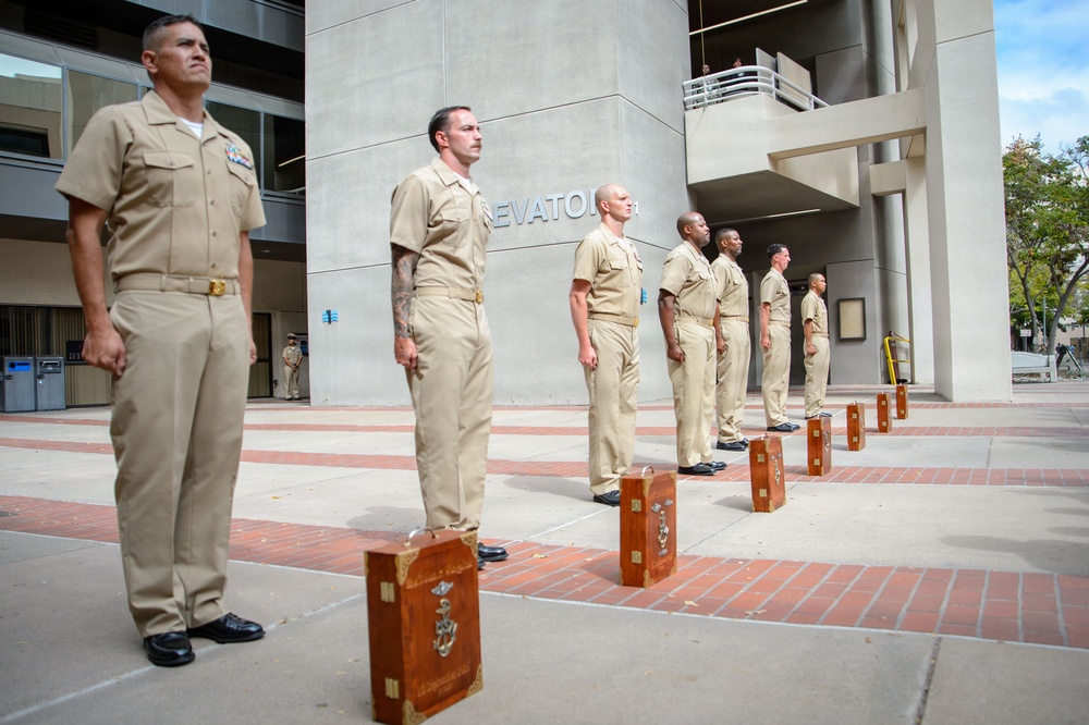 A group of U.S. Navy personnel in khaki service uniforms stand at attention in a formal outdoor ceremony, likely a promotion or retirement event. Each sailor stands beside a wooden presentation box, which typically contains a ceremonial shadow box or award case. The setting appears to be a military installation or government building plaza.