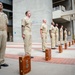 A group of U.S. Navy personnel in khaki service uniforms stand at attention in a formal outdoor ceremony, likely a promotion or retirement event. Each sailor stands beside a wooden presentation box, which typically contains a ceremonial shadow box or award case. The setting appears to be a military installation or government building plaza.