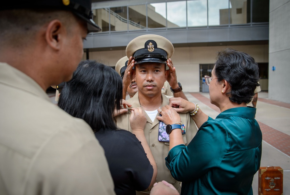 A U.S. Navy Chief Petty Officer promotion ceremony is taking place outdoors near a modern building. Family members or colleagues are pinning insignia onto the newly promoted sailor's uniform collar while another person adjusts his Chief Petty Officer cover (hat). This is a traditional 'pinning ceremony' marking the sailor's advancement to the Chief Petty Officer rank.