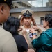 A U.S. Navy Chief Petty Officer promotion ceremony is taking place outdoors near a modern building. Family members or colleagues are pinning insignia onto the newly promoted sailor's uniform collar while another person adjusts his Chief Petty Officer cover (hat). This is a traditional 'pinning ceremony' marking the sailor's advancement to the Chief Petty Officer rank.