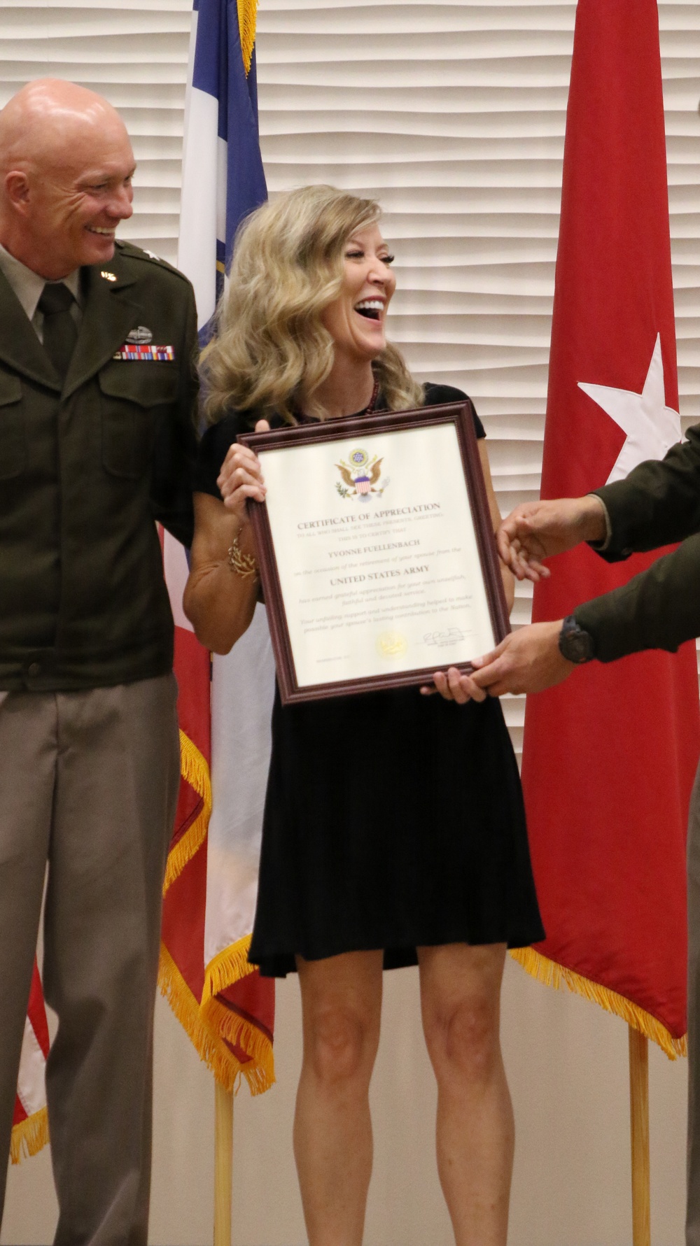 A woman in a black dress is joyfully receiving a framed United States Army Certificate of Appreciation, likely awarded to 'Yvonne Fellenbach' based on visible text. She is flanked by a smiling military officer in Army green uniform on her left and another uniformed individual presenting the certificate on her right. The ceremony takes place in front of military flags, including a general officer's red flag with a white star.