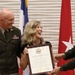 A woman in a black dress is joyfully receiving a framed United States Army Certificate of Appreciation, likely awarded to 'Yvonne Fellenbach' based on visible text. She is flanked by a smiling military officer in Army green uniform on her left and another uniformed individual presenting the certificate on her right. The ceremony takes place in front of military flags, including a general officer's red flag with a white star.