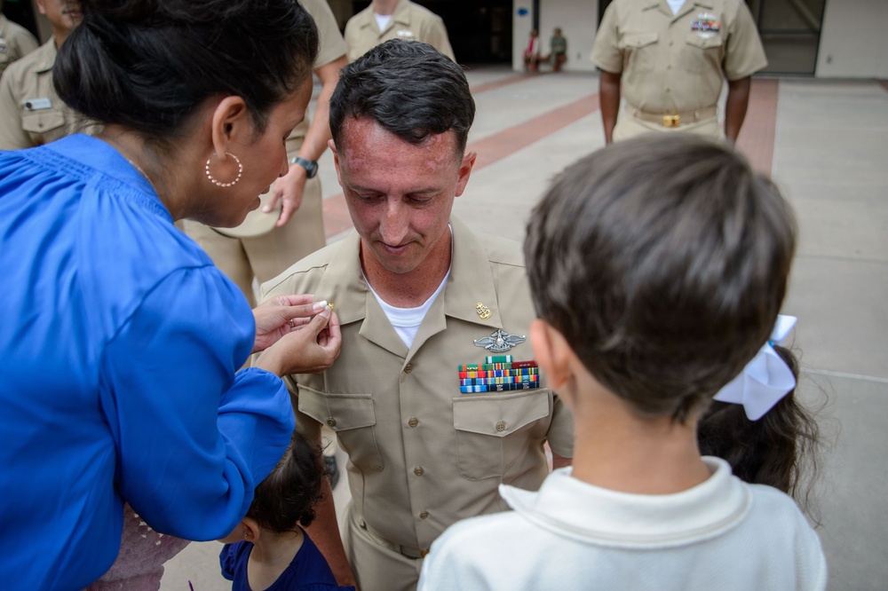 A U.S. Navy promotion ceremony is taking place outdoors, where a woman in a blue top is pinning a new rank insignia onto a male sailor's uniform collar. The sailor is wearing a tan/khaki Navy uniform adorned with multiple service ribbons and a warfare qualification badge. Children are present, suggesting this is a family-attended military promotion event.