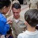 A U.S. Navy promotion ceremony is taking place outdoors, where a woman in a blue top is pinning a new rank insignia onto a male sailor's uniform collar. The sailor is wearing a tan/khaki Navy uniform adorned with multiple service ribbons and a warfare qualification badge. Children are present, suggesting this is a family-attended military promotion event.