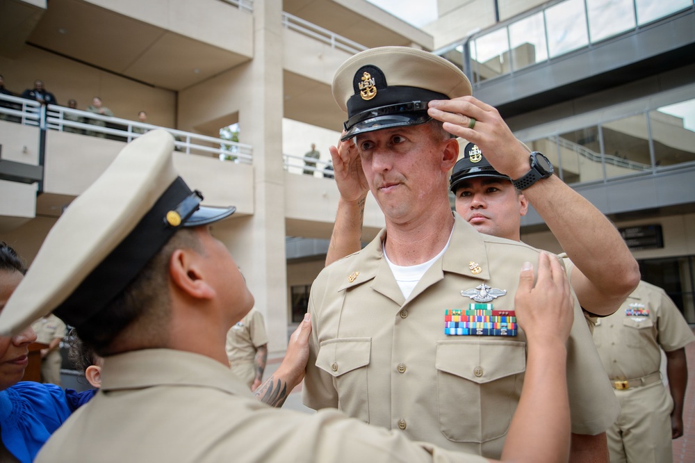 A U.S. Navy promotion or pinning ceremony is taking place outdoors in what appears to be a military installation courtyard. A senior enlisted sailor is having his combination cover (cap) placed on his head, while another person adjusts insignia on his uniform. The central figure wears a khaki service uniform with multiple ribbons and a warfare device, indicating significant service experience.