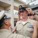 A U.S. Navy promotion or pinning ceremony is taking place outdoors in what appears to be a military installation courtyard. A senior enlisted sailor is having his combination cover (cap) placed on his head, while another person adjusts insignia on his uniform. The central figure wears a khaki service uniform with multiple ribbons and a warfare device, indicating significant service experience.