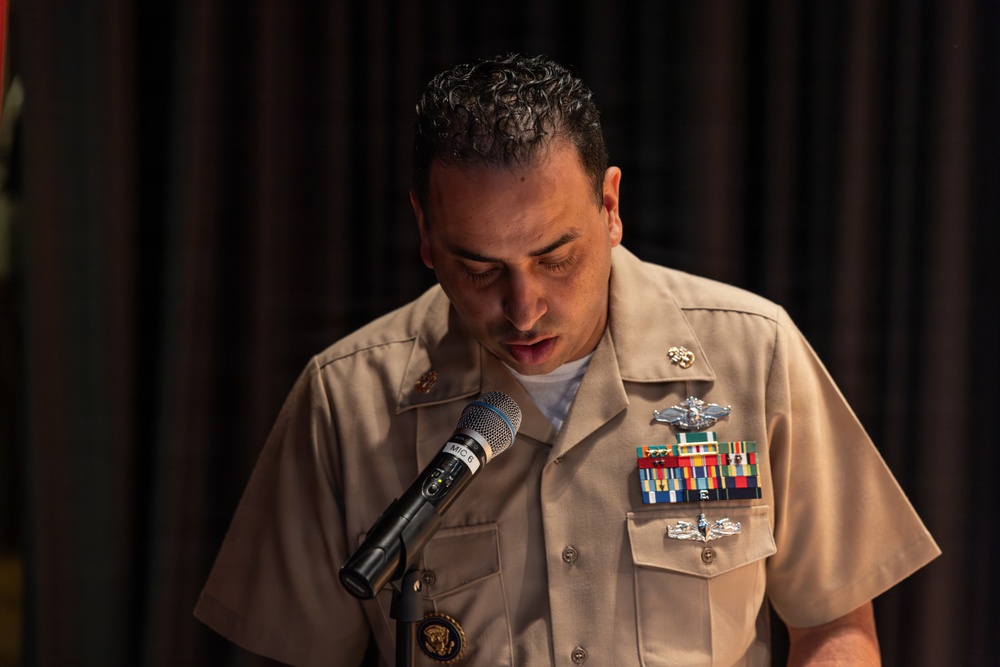 A U.S. Navy officer in tan service uniform is speaking at a microphone, looking downward, likely reading from notes or a script. The officer displays multiple ribbons, warfare qualification badges, and insignia on their uniform. The setting appears to be an indoor formal or ceremonial event with dark curtain backdrop.