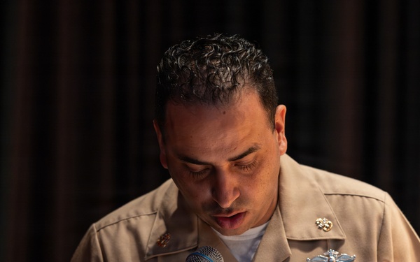A U.S. Navy officer in tan service uniform is speaking at a microphone, looking downward, likely reading from notes or a script. The officer displays multiple ribbons, warfare qualification badges, and insignia on their uniform. The setting appears to be an indoor formal or ceremonial event with dark curtain backdrop.