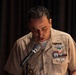 A U.S. Navy officer in tan service uniform is speaking at a microphone, looking downward, likely reading from notes or a script. The officer displays multiple ribbons, warfare qualification badges, and insignia on their uniform. The setting appears to be an indoor formal or ceremonial event with dark curtain backdrop.