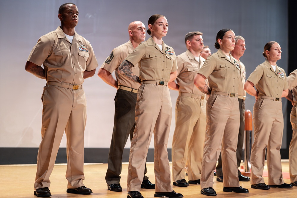 A group of military personnel in tan/khaki service uniforms are standing at attention or parade rest on a wooden stage or platform. The individuals display various rank insignia, ribbons, and badges on their uniforms. The formation appears to be part of a formal military ceremony or official event.