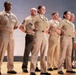 A group of military personnel in tan/khaki service uniforms are standing at attention or parade rest on a wooden stage or platform. The individuals display various rank insignia, ribbons, and badges on their uniforms. The formation appears to be part of a formal military ceremony or official event.