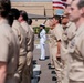 A U.S. Navy military ceremony or formation is taking place outdoors on a paved area, likely at a naval base. The majority of personnel are wearing tan/khaki Navy working uniforms, while one sailor in the center stands out in dress whites and holds a rifle at attention. An American flag with gold fringe is visible being carried in the background.