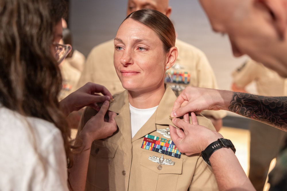 A U.S. Navy service member in Service Khaki uniform is undergoing a promotion or pinning ceremony. Two individuals — one on each side — are simultaneously pinning new rank insignia onto the collar of her uniform. The subject appears emotional, with a restrained smile, suggesting personal significance of the moment.