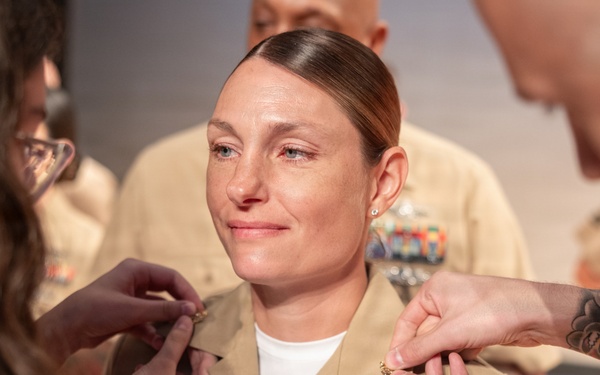 A U.S. Navy service member in Service Khaki uniform is undergoing a promotion or pinning ceremony. Two individuals — one on each side — are simultaneously pinning new rank insignia onto the collar of her uniform. The subject appears emotional, with a restrained smile, suggesting personal significance of the moment.