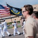 A United States Navy color guard ceremony is taking place on a paved outdoor area, likely at a naval installation. Personnel in white dress uniforms are carrying the American flag and the United States Navy flag while other sailors in khaki/tan uniforms stand at attention observing. The scene appears to be a formal military parade or change of command ceremony.